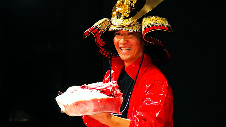 The host in a samurai helmet holding a block of tuna belly